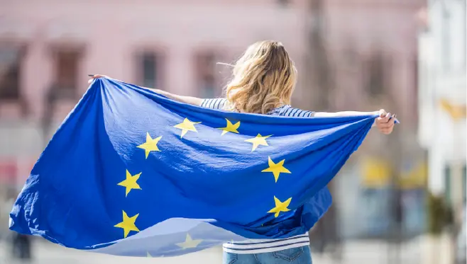 Młoda kobieta odwrócona tyłem do aparatu trzymająca za plecami powiewającą Flagę Europejską