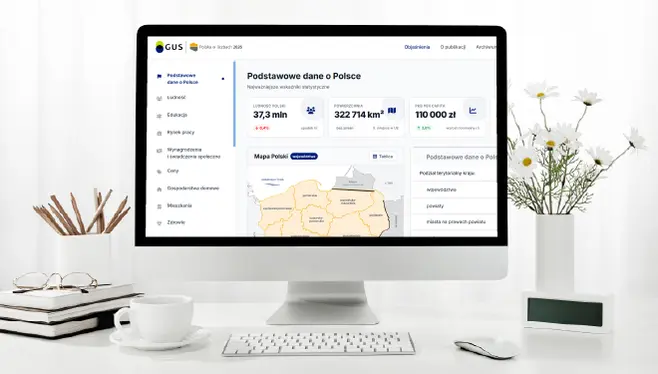 Monitor komputera stoi na biurku. Obok niego kubek z ołówkami, doniczka z białymi kwiatami, myszka, klawiatura i notatnik. Na ekranie strona GUS Polska w liczbach