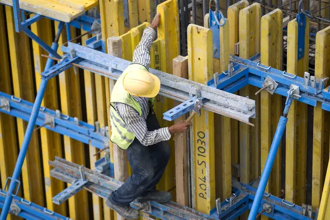 A man in work clothes and a helmet is installing a yellow shuttering panel on a construction site