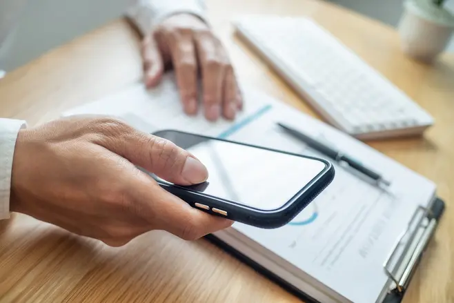 Osoba analizująca dane. Widać dłonie trzymające smartphone oraz notatki i klawiaturę na biurku