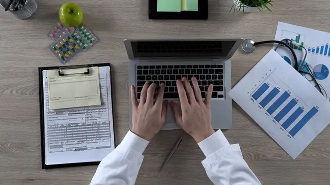 A top-down view of a man’s hands, clad in a long-sleeved shirt, typing on a laptop keyboard. Next to the laptop on the desk are scattered sheets of paper with charts and notes