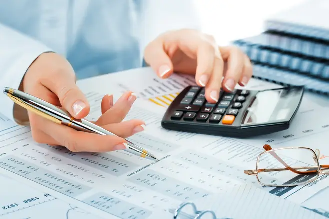A woman analysing data. She is holding a pen in one hand and has her other hand resting on a calculator; there are documents spread out on her desk