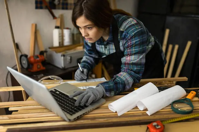 Kobieta w warsztacie stolarskim, ubrana w odzież roboczą, patrzy na ekran laptopa