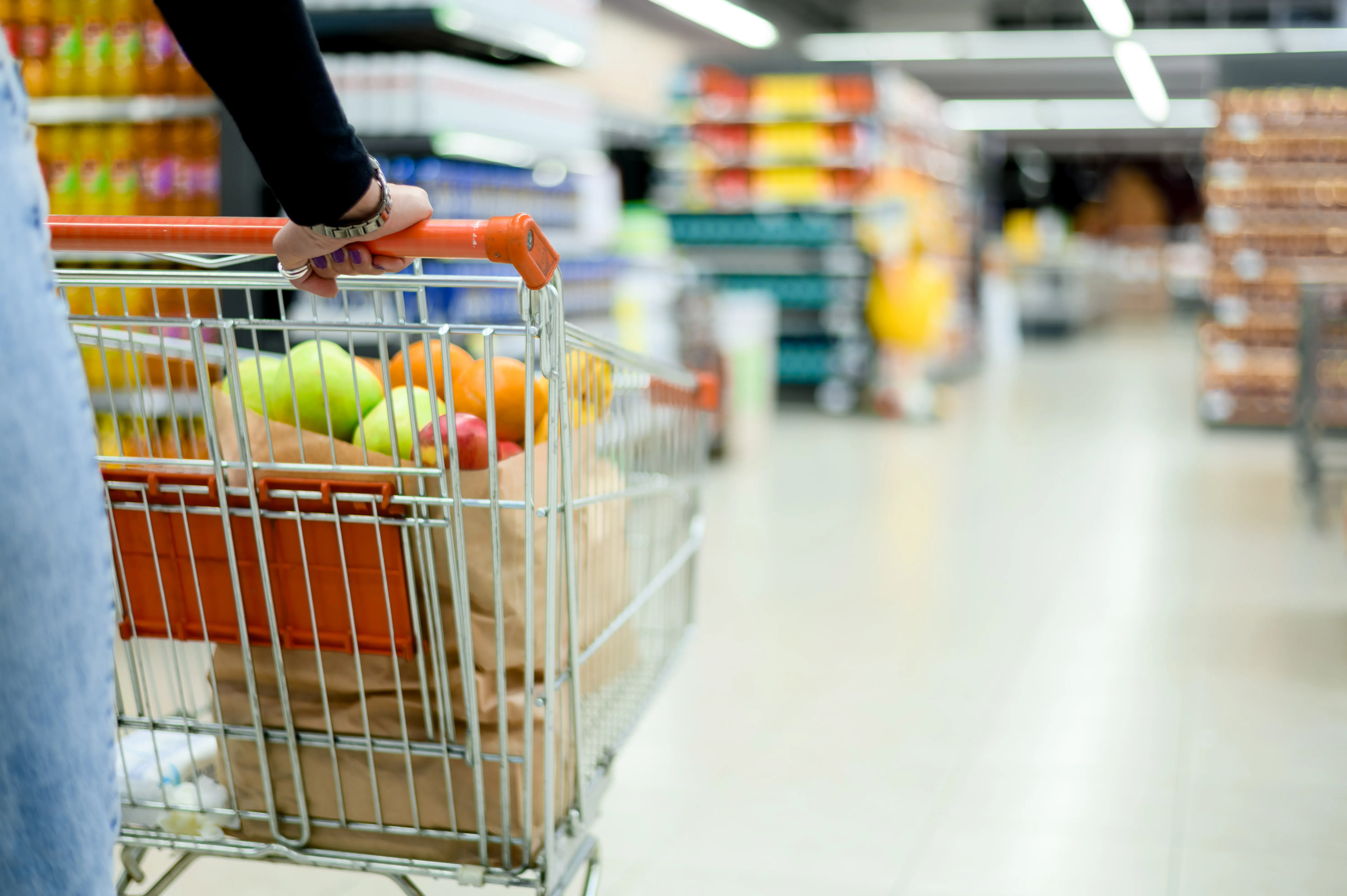 Na grafice znajduje się wózek sklepowy z towarami spożywczymi na tle supermarketu