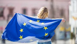 Powiększ obraz: Młoda kobieta odwrócona tyłem do aparatu trzymająca za plecami powiewającą Flagę Europejską